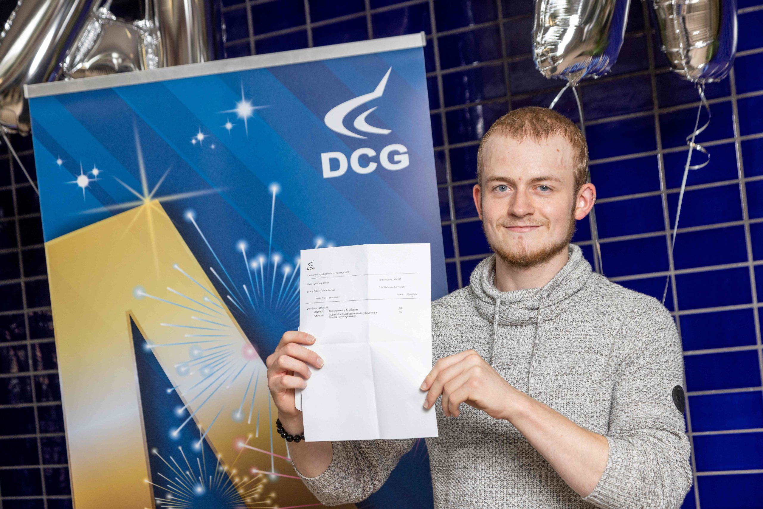 Young male with short blonde hair wearing a grey jumper holding a document with exam results.